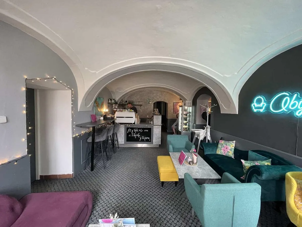 Interiér bistra Obývák — klenutý strop, bar, tyrkysová a růžová sofa a neonový nápis Obývák